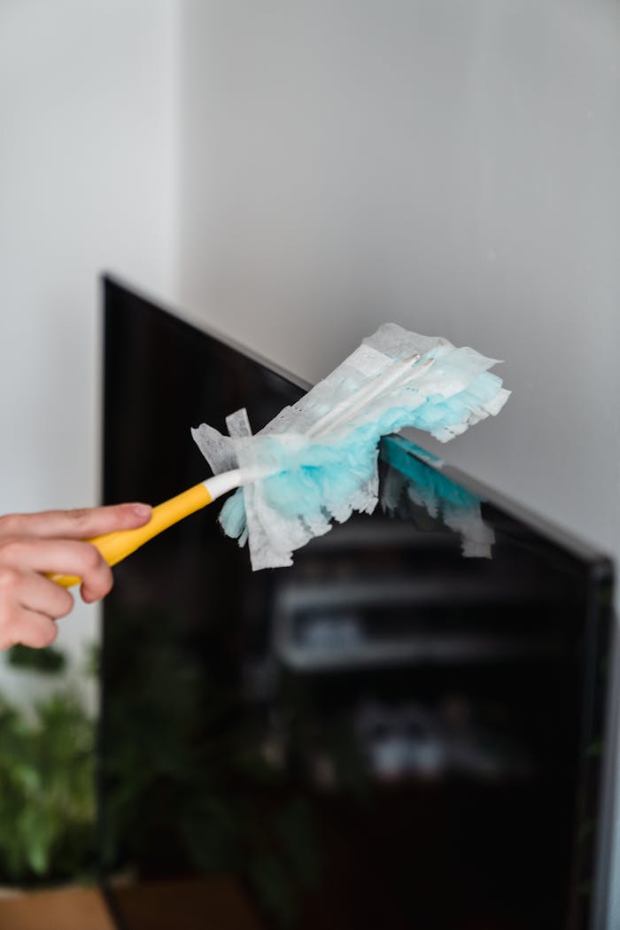 Close-up of a hand dusting a TV screen with a blue feather duster indoors.